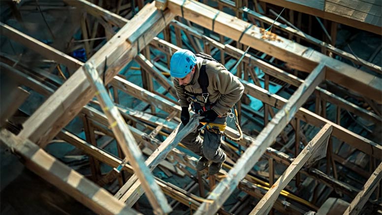 Construction worker wearing full-body harnesses and high-vis gear, tethered on a raised platform with safety lines.