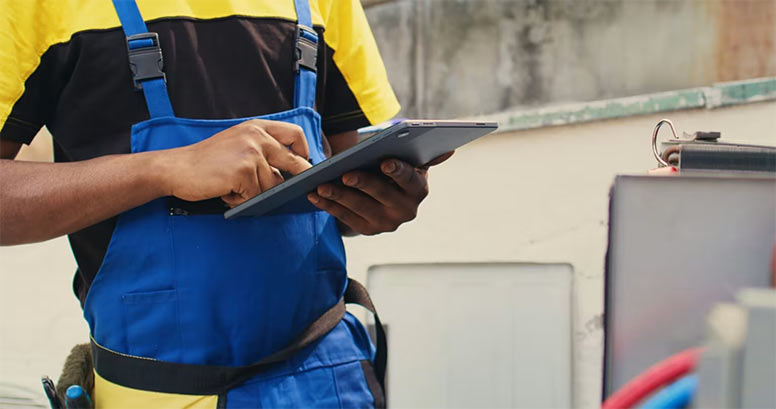 Close-up of an HVAC technician in blue overalls using a tablet to log data during an outdoor air conditioning unit inspection