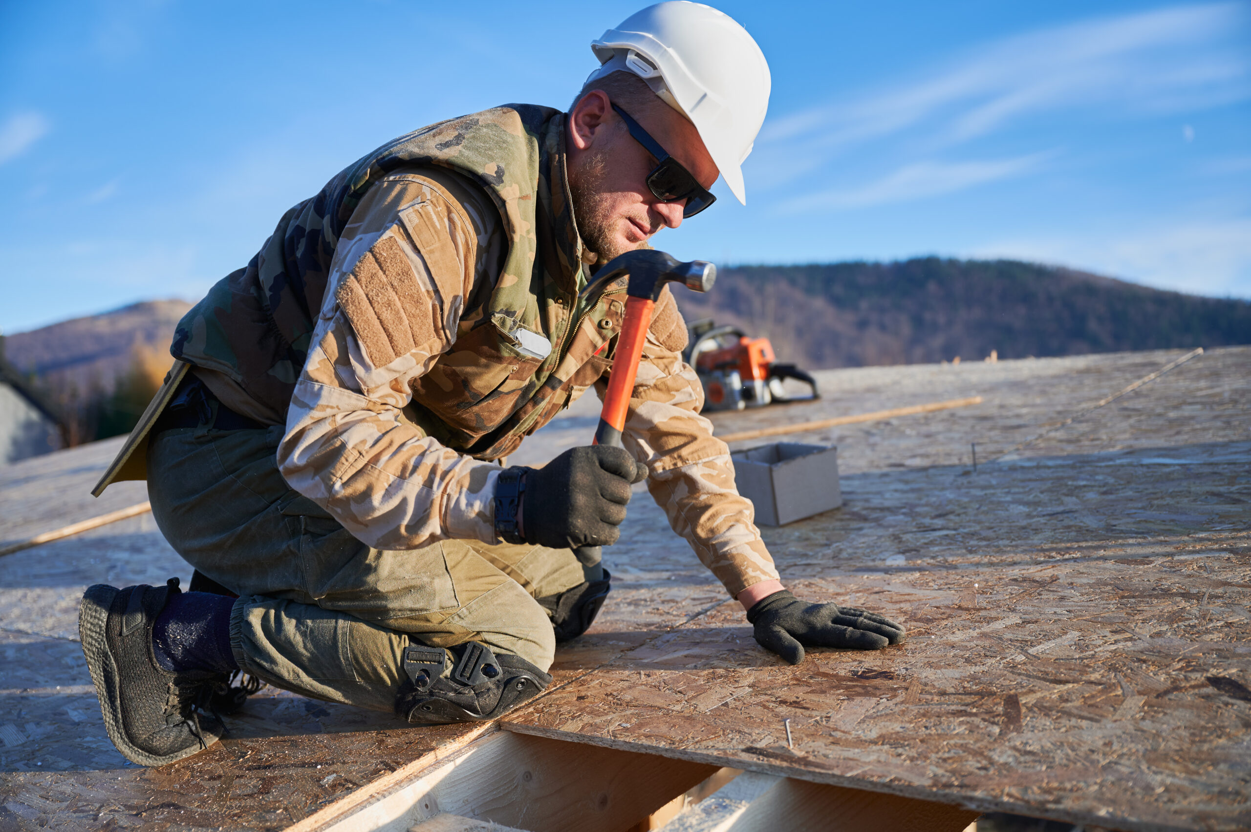 Roofer hammering nail into OSB panel on the roof top of future cottage. Roofing worker building wooden frame house. Roofing and construction concept.