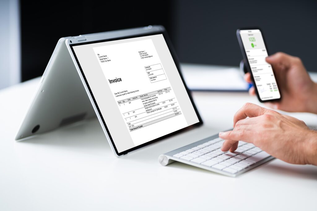 Person typing on keyboard while viewing an invoice on a tablet and smartphone screen.