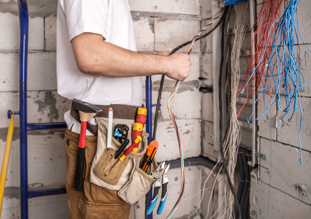 Electrician with a full tool belt holding electrical wires in front of a panel with exposed cables.