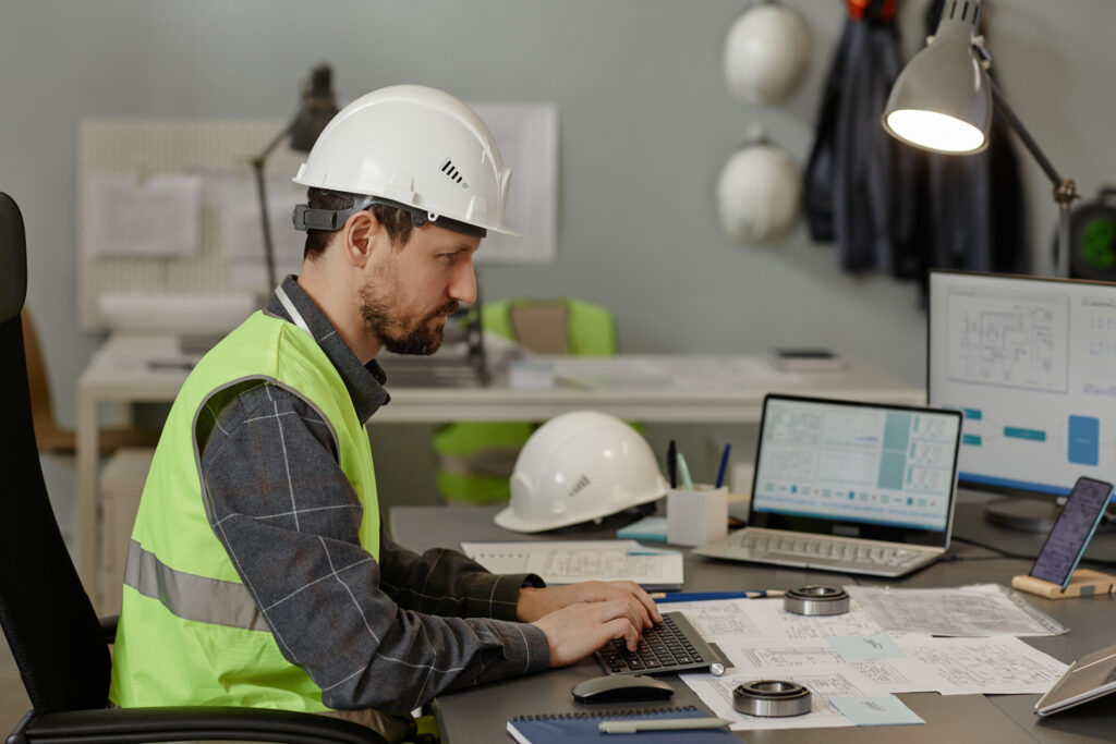 Engineer wearing hardhat at workplace in office and using multiple computer devices for calculation and design