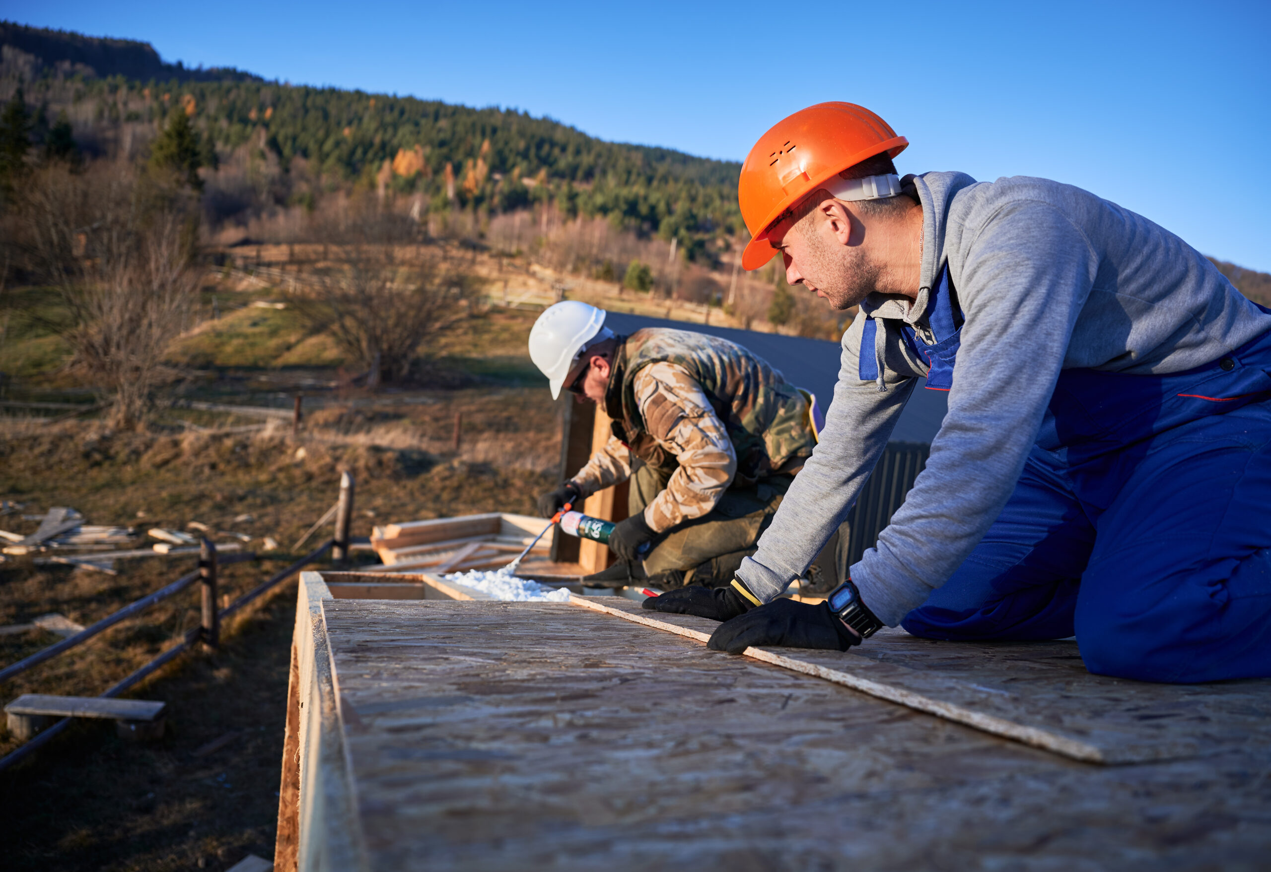 Roofing builder doing thermal insulation on roof of wooden frame house. Roofing worker spraying polyurethane foam on rooftop of future cottage. Construction and insulation concept.