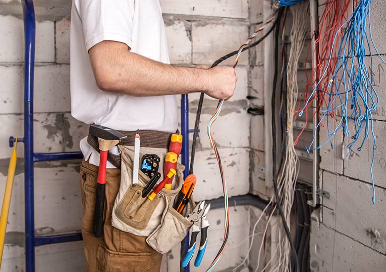 Electrician with a full tool belt holding electrical wires in front of a panel with exposed cables.