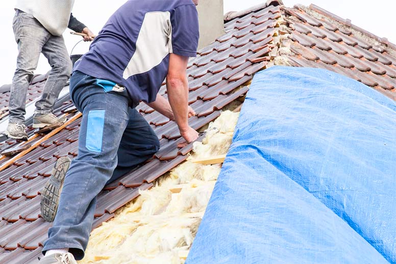 Two roofers work on installing red ceramic roof tiles while insulation and wooden supports are visible beneath a blue tarp.