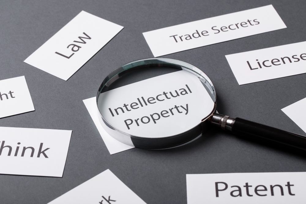Magnifying glass over words “Intellectual Property” with related terms like Patent and Trade Secrets, representing intangible assets.