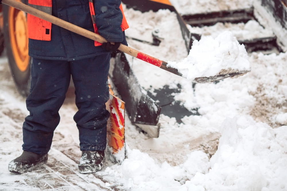 Worker shoveling snow manually next to snow removal machinery, illustrating DIY vs professional service comparison.